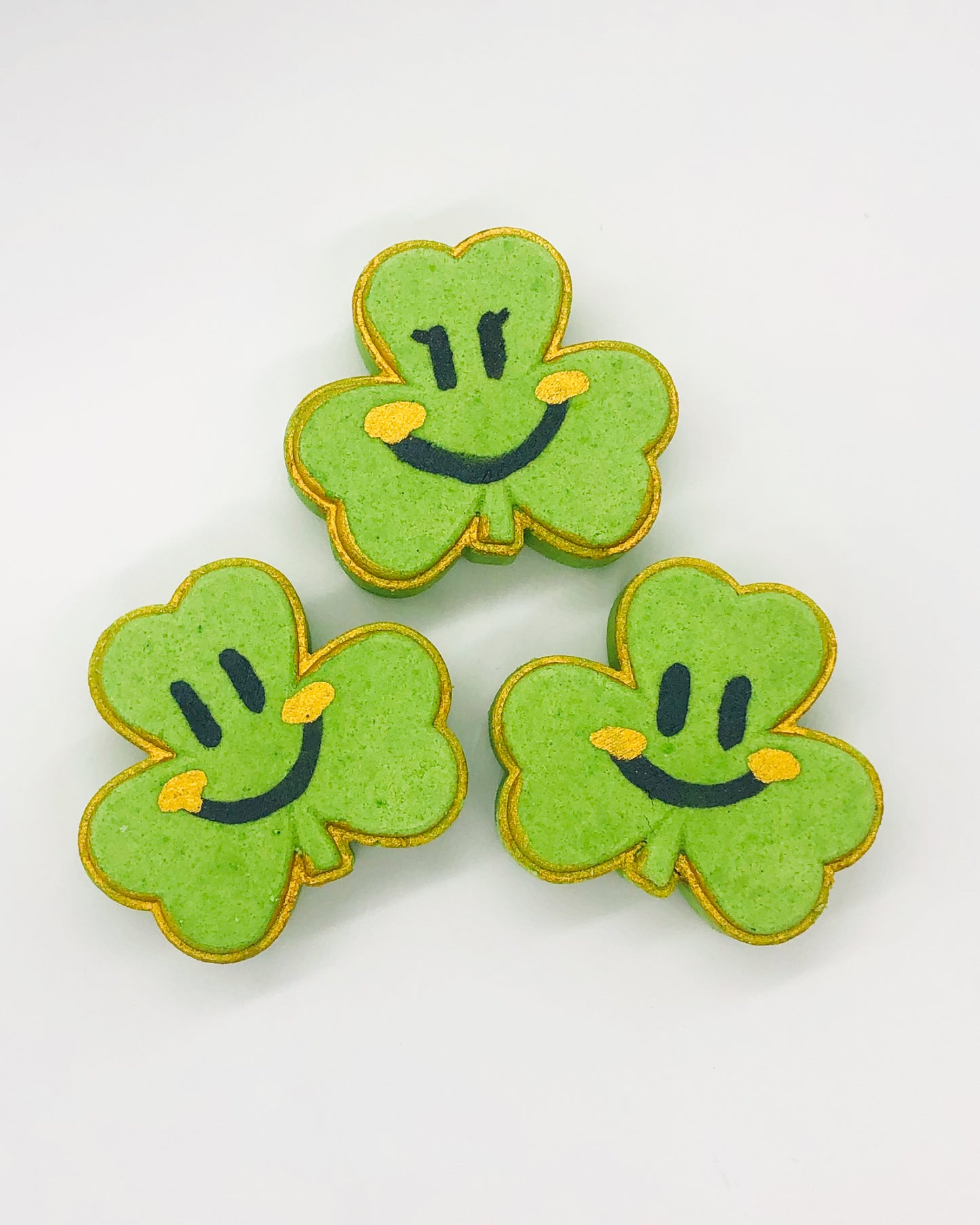 Three green shamrock-shaped bath bombs with a smiling face drawn on each, against a white background.