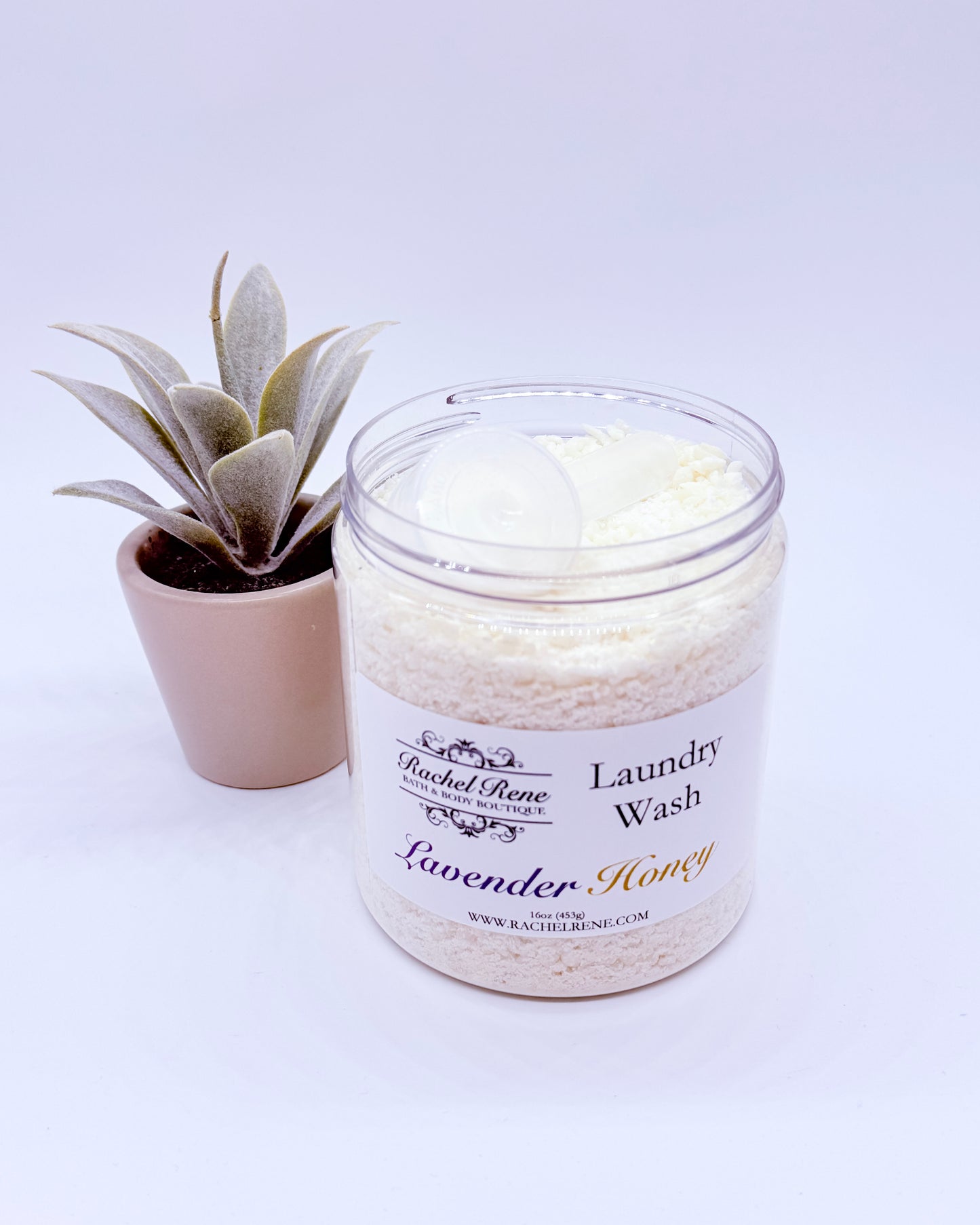 An open jar of Rachel Rene Lavender Honey laundry wash with a small scoop  with a plant on a white background