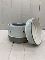 A circular ceramic wax warmer. It is light gray with a band of dark gray on the bottom. There are two holes where light can shine through. The top cup is detached and leaning against the base.