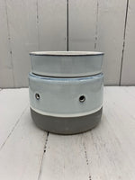 A circular ceramic wax warmer. It is light gray with a band of dark gray on the bottom. There are two holes where light can shine through.